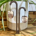 Wooden dangle earrings with a mountain landscape engraving, displayed on a wooden stand in front of a ceramic planter with green plant leaves.