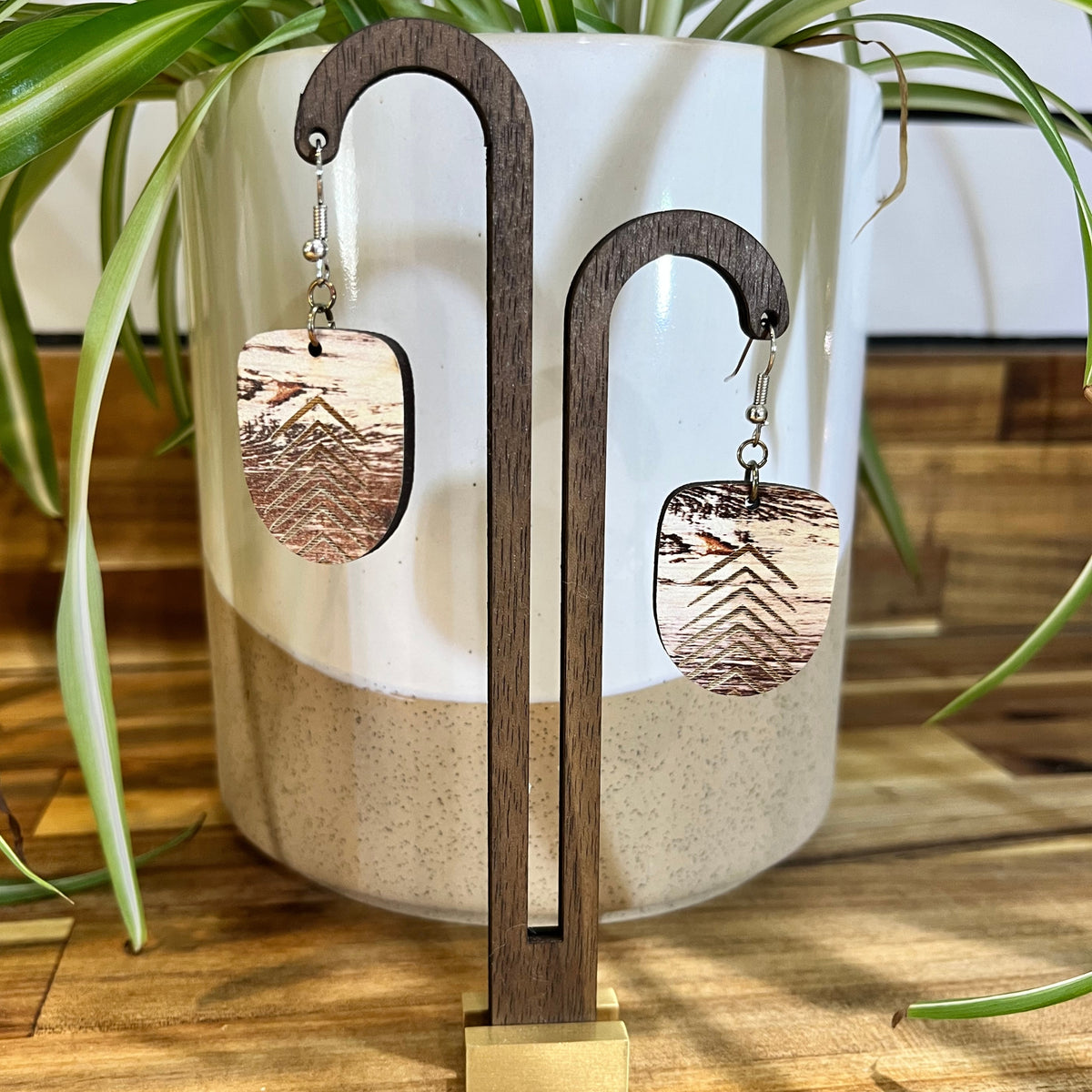 Wooden dangle earrings with a mountain landscape engraving, displayed on a wooden stand in front of a ceramic planter with green plant leaves.
