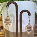 A pair of dangling earrings displayed on a wooden stand, featuring diamond-shaped wooden plates with horizontal cutout lines and silver-toned fishhook wires, set against a white ceramic planter with green leaves.