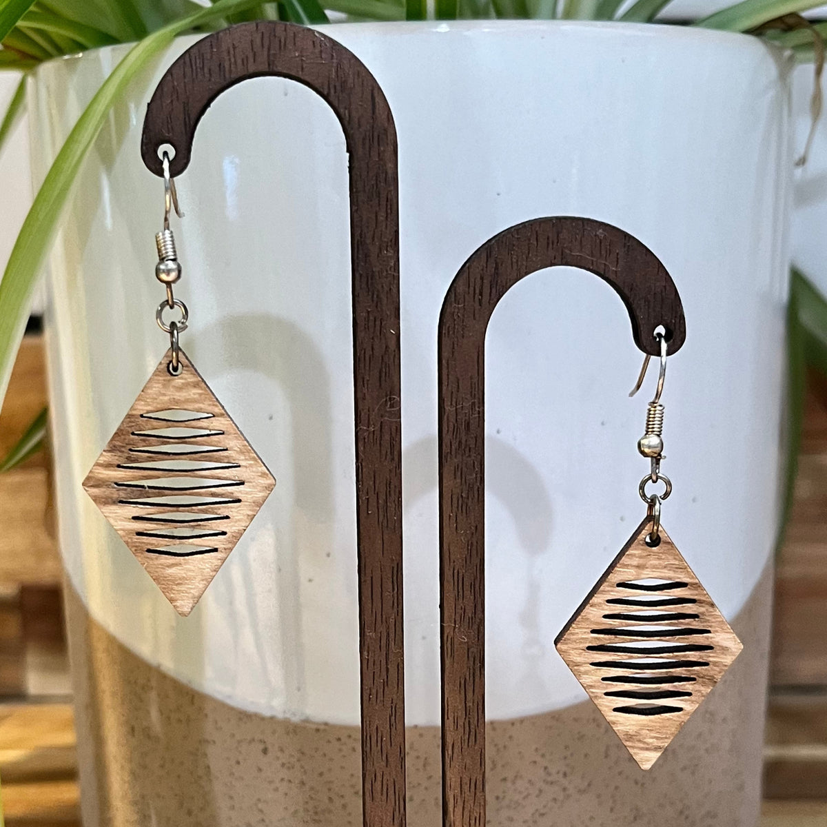 A pair of dangling earrings displayed on a wooden stand, featuring diamond-shaped wooden plates with horizontal cutout lines and silver-toned fishhook wires, set against a white ceramic planter with green leaves.