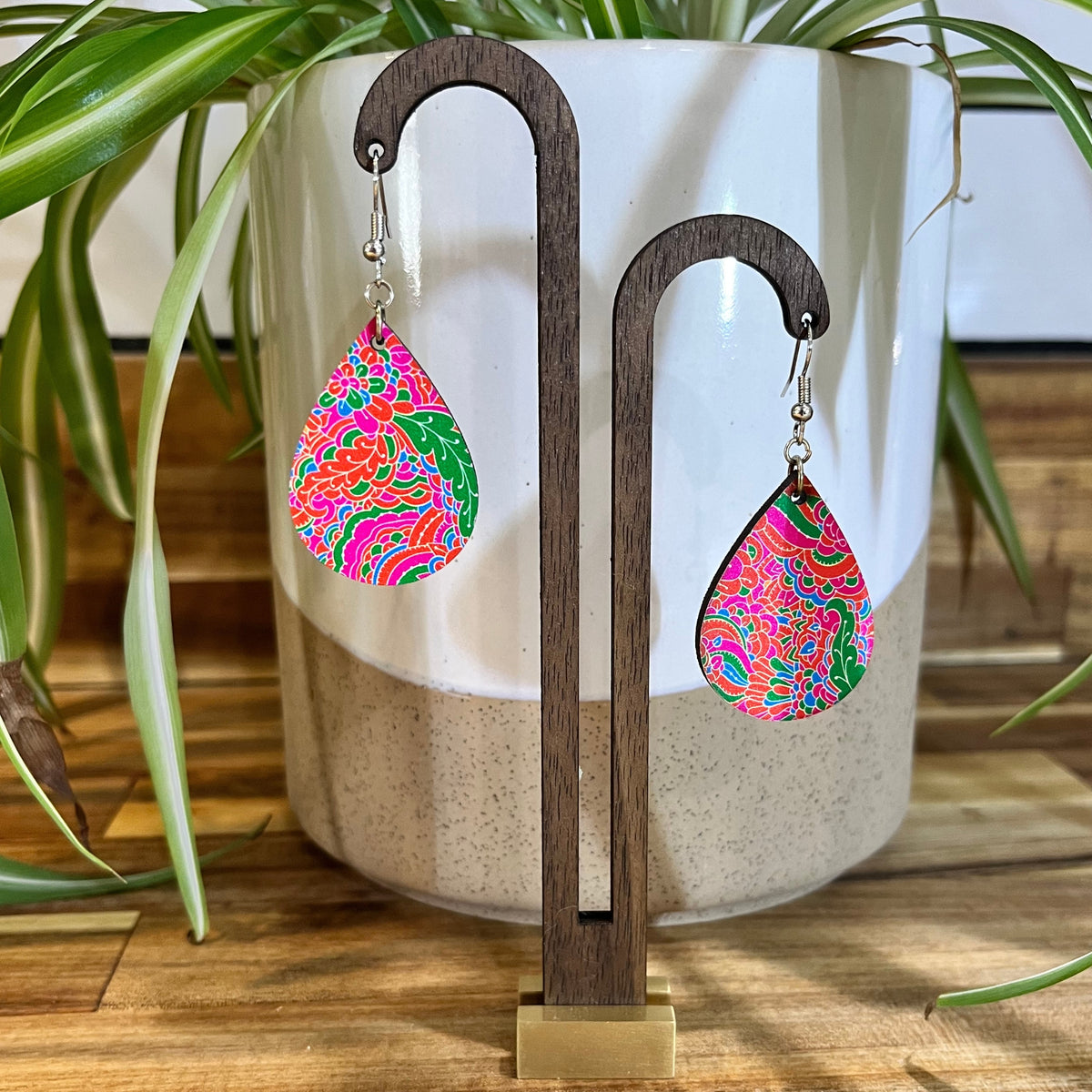 A pair of colorful teardrop-shaped dangle earrings displayed on a dark wooden stand in front of a white ceramic planter with green plant leaves.