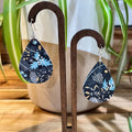 Teardrop-shaped dangle earrings with a navy blue botanical pattern featuring light blue, white, and gold details, displayed on a wooden stand in front of a potted plant.