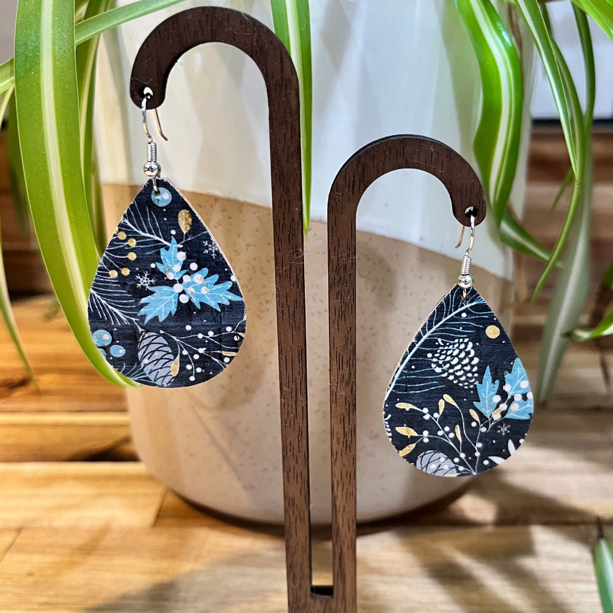 Teardrop-shaped dangle earrings with a navy blue botanical pattern featuring light blue, white, and gold details, displayed on a wooden stand in front of a potted plant.