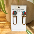 A pair of drop earrings displayed on a white card from Willis Design Co. The earrings feature round, colorful top studs with a red, blue, and black marbled pattern, connected by gold-tone wires to elongated, open-top rectangular dangles in a gold-tone finish.