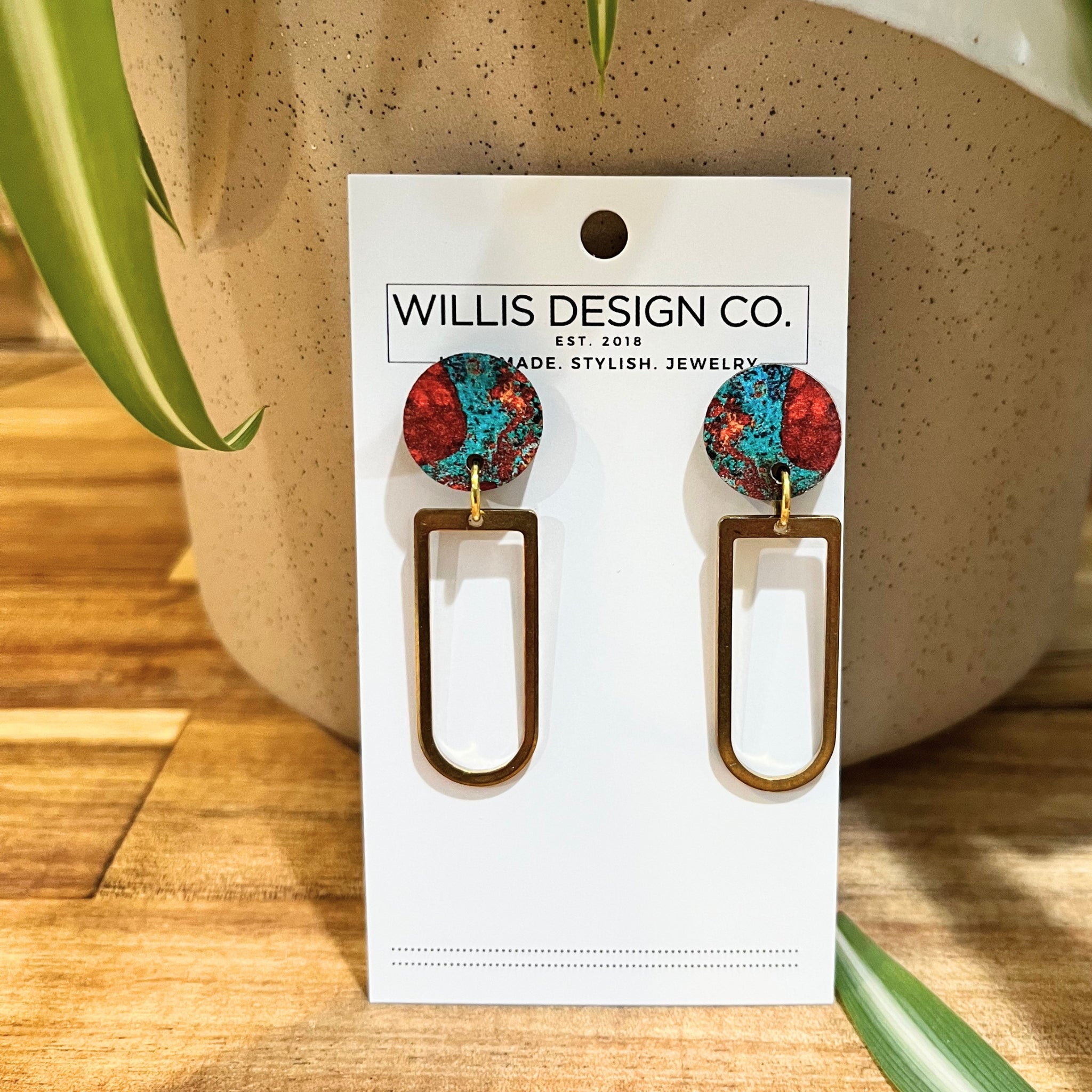 A pair of drop earrings displayed on a white card from Willis Design Co. The earrings feature round, colorful top studs with a red, blue, and black marbled pattern, connected by gold-tone wires to elongated, open-top rectangular dangles in a gold-tone finish.