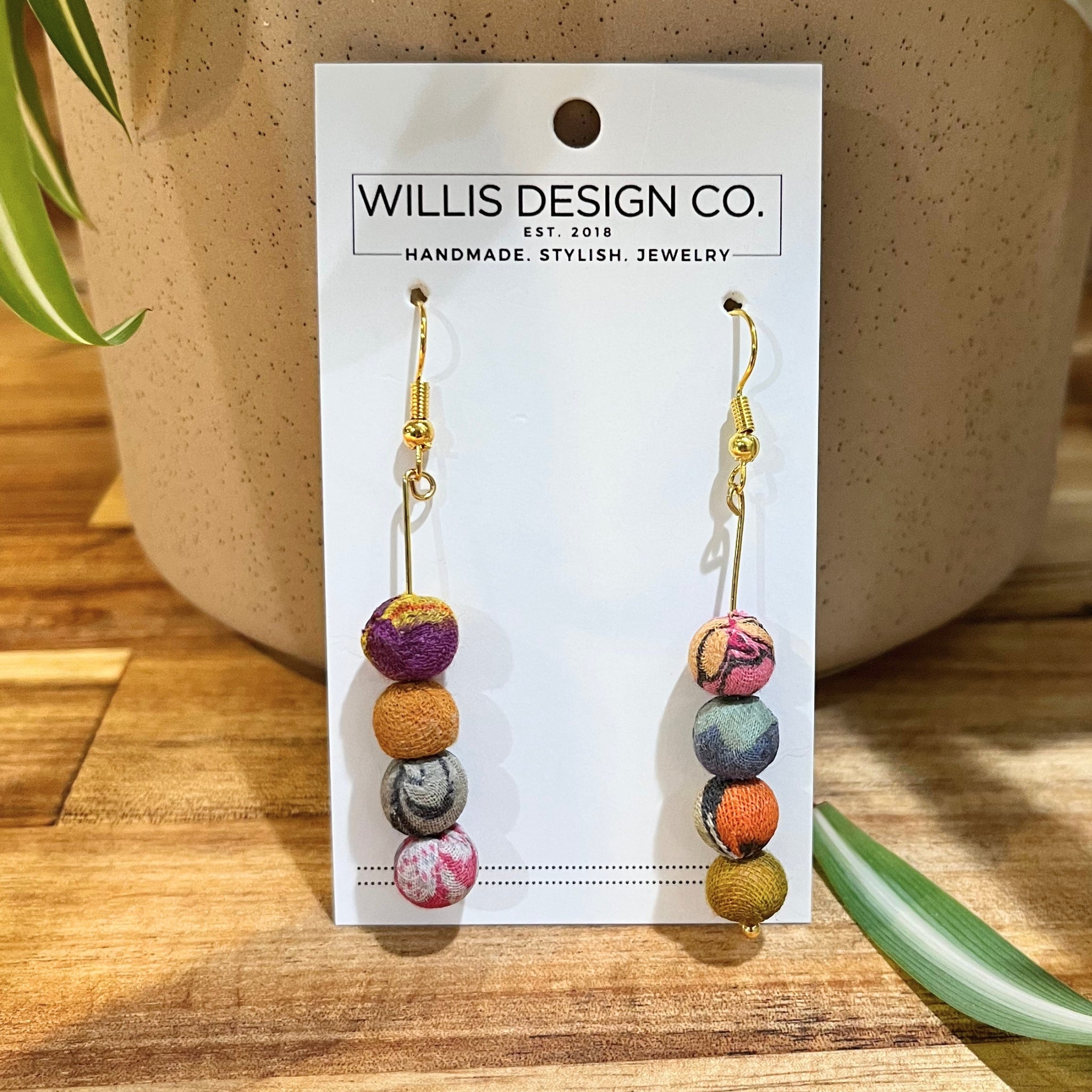 A pair of handmade fabric ball dangle earrings by Willis Design Co. displayed on a white brand card.