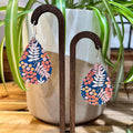Teardrop-shaped dangle earrings with a navy blue background and a colorful botanical pattern of pink, orange, and white leaves and berries, displayed on dark wooden hangers in front of a potted plant.