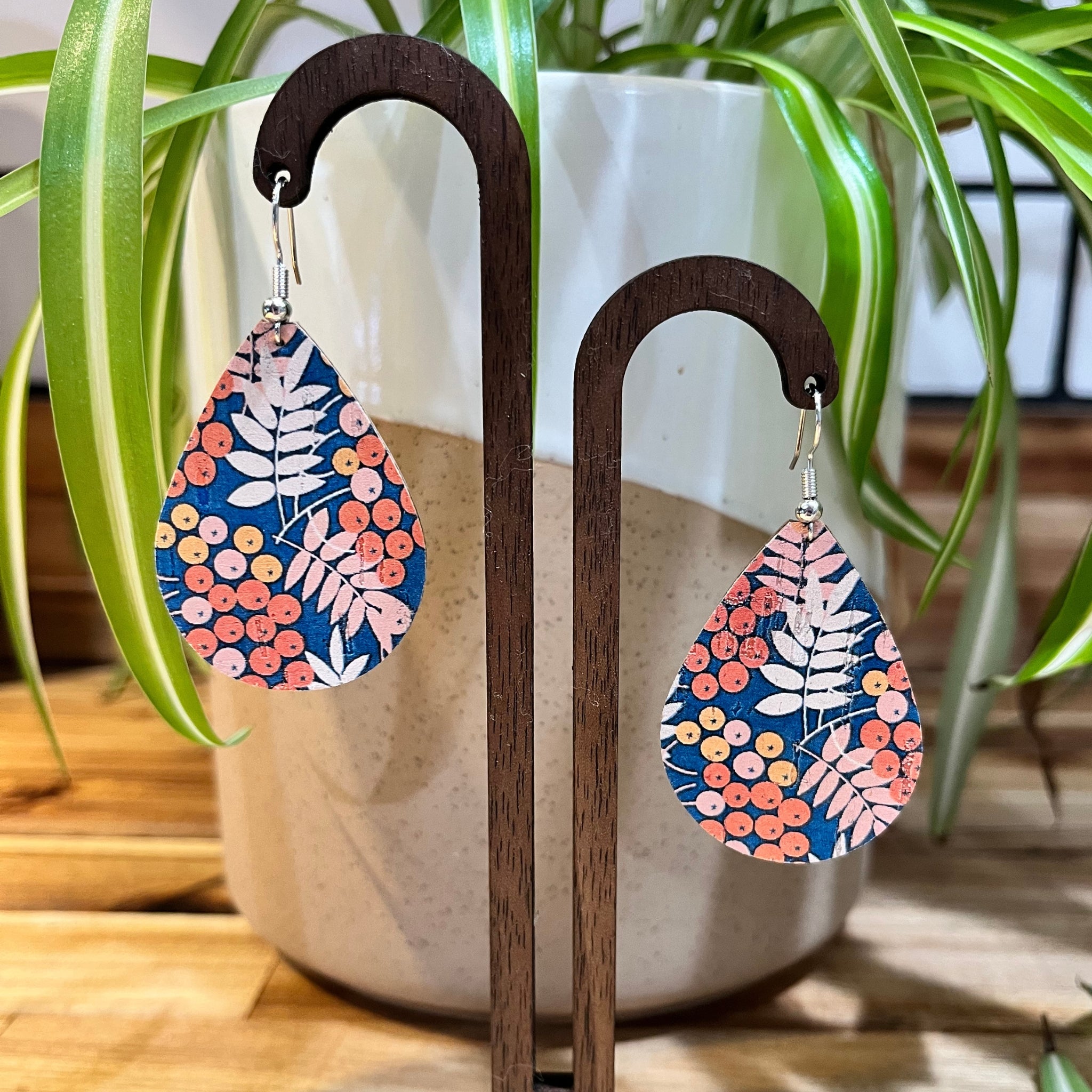 Teardrop-shaped dangle earrings with a navy blue background and a colorful botanical pattern of pink, orange, and white leaves and berries, displayed on dark wooden hangers in front of a potted plant.