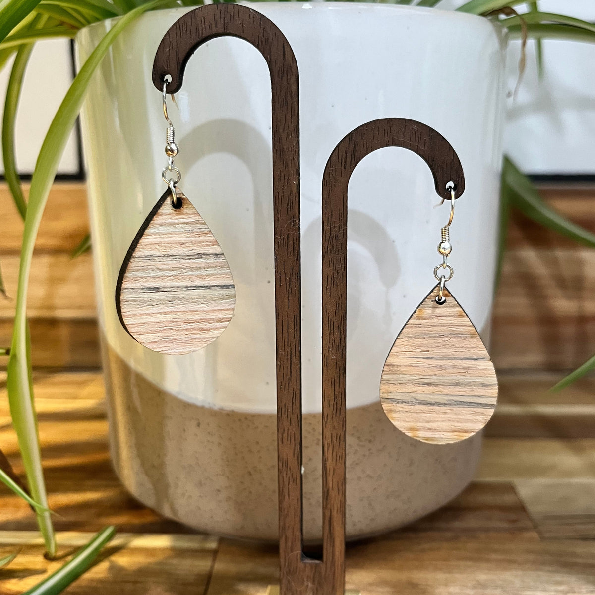 Teardrop-shaped wooden dangle earrings with a natural wood grain pattern, displayed on a dark wood stand in front of a ceramic planter with green plant leaves.