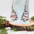 A pair of dangling earrings shaped like Christmas trees, displayed on a white card stand. The trees are white with red stripes and black polka dots, hanging from silver fishhook wires.