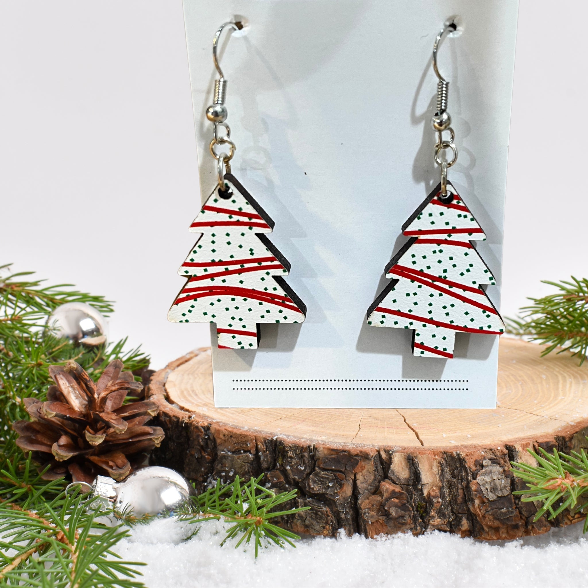 A pair of dangling earrings shaped like Christmas trees, displayed on a white card stand. The trees are white with red stripes and black polka dots, hanging from silver fishhook wires.