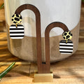 A pair of geometric dangle earrings displayed on a wooden stand. The earrings feature a round black-and-white striped square drop, a round yellow-and-black cow-print circle, and a small gold-tone connector.