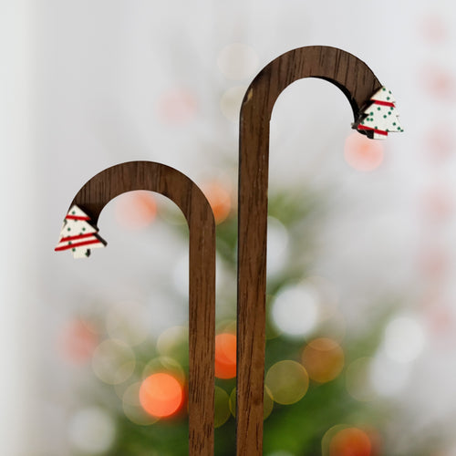 Two curved wooden decorative stakes with small Christmas tree ornaments at the tips, shown against a softly blurred festive background with lights.