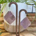Two diamond-shaped dangle earrings with a textured, speckled dusty purple surface, displayed on a wooden stand in front of a white planter with green spider plants.