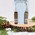 A pair of rectangular drop earrings with a dark wood grain top half and a white geometric patterned bottom half, hanging from silver hooks on a light blue display card. The card is labeled 'WILLIS DESIGN CO.