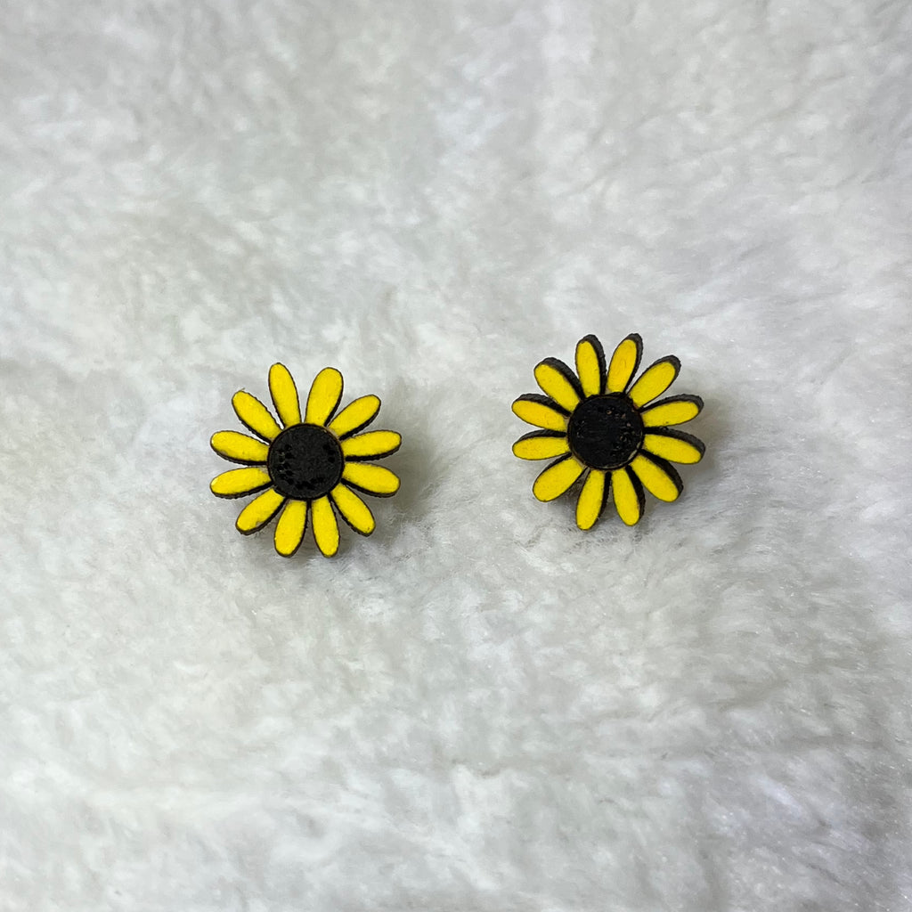 A pair of small daisy-shaped stud earrings with bright yellow petals, black centers, and thin dark outlines, placed on a white textured fabric background.