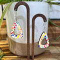 A pair of dangling triangle earrings with a colorful polka-dot pattern on a white background, displayed on a wooden stand in front of a white ceramic planter.