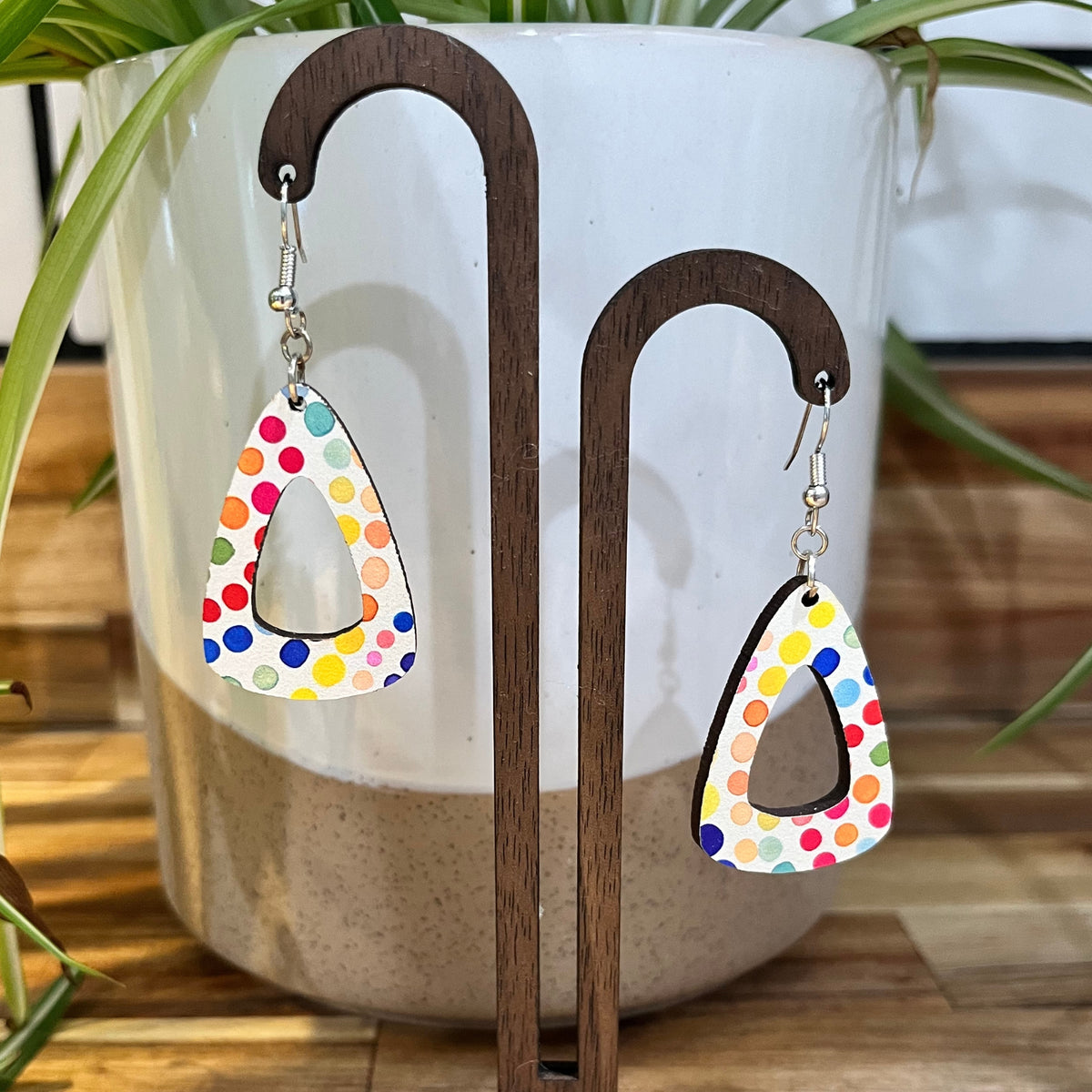 A pair of dangling triangle earrings with a colorful polka-dot pattern on a white background, displayed on a wooden stand in front of a white ceramic planter.