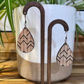 Teardrop-shaped wooden dangle earrings with a black and beige chevron pattern, displayed on dark wood stands in front of a white ceramic pot with green plant leaves.