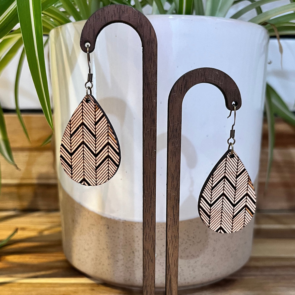 Teardrop-shaped wooden dangle earrings with a black and beige chevron pattern, displayed on dark wood stands in front of a white ceramic pot with green plant leaves.