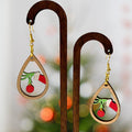 Teardrop-shaped wooden dangle earrings with a Christmas tree branch design featuring a green hand holding a red ornament, displayed on a wooden stand against a blurred festive background.