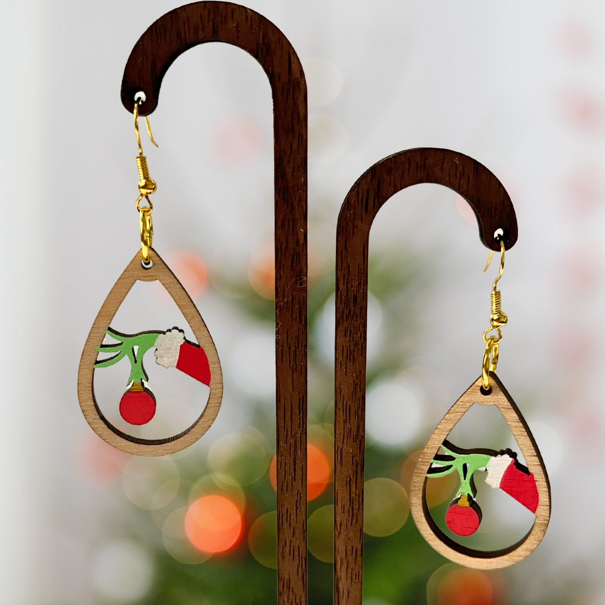 Teardrop-shaped wooden dangle earrings with a Christmas tree branch design featuring a green hand holding a red ornament, displayed on a wooden stand against a blurred festive background.