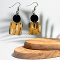 A pair of geometric drop earrings with black circular discs and square gold-toned charms featuring black and white tribal-style patterns, displayed on a wooden stand against a white background with soft leaf shadows.