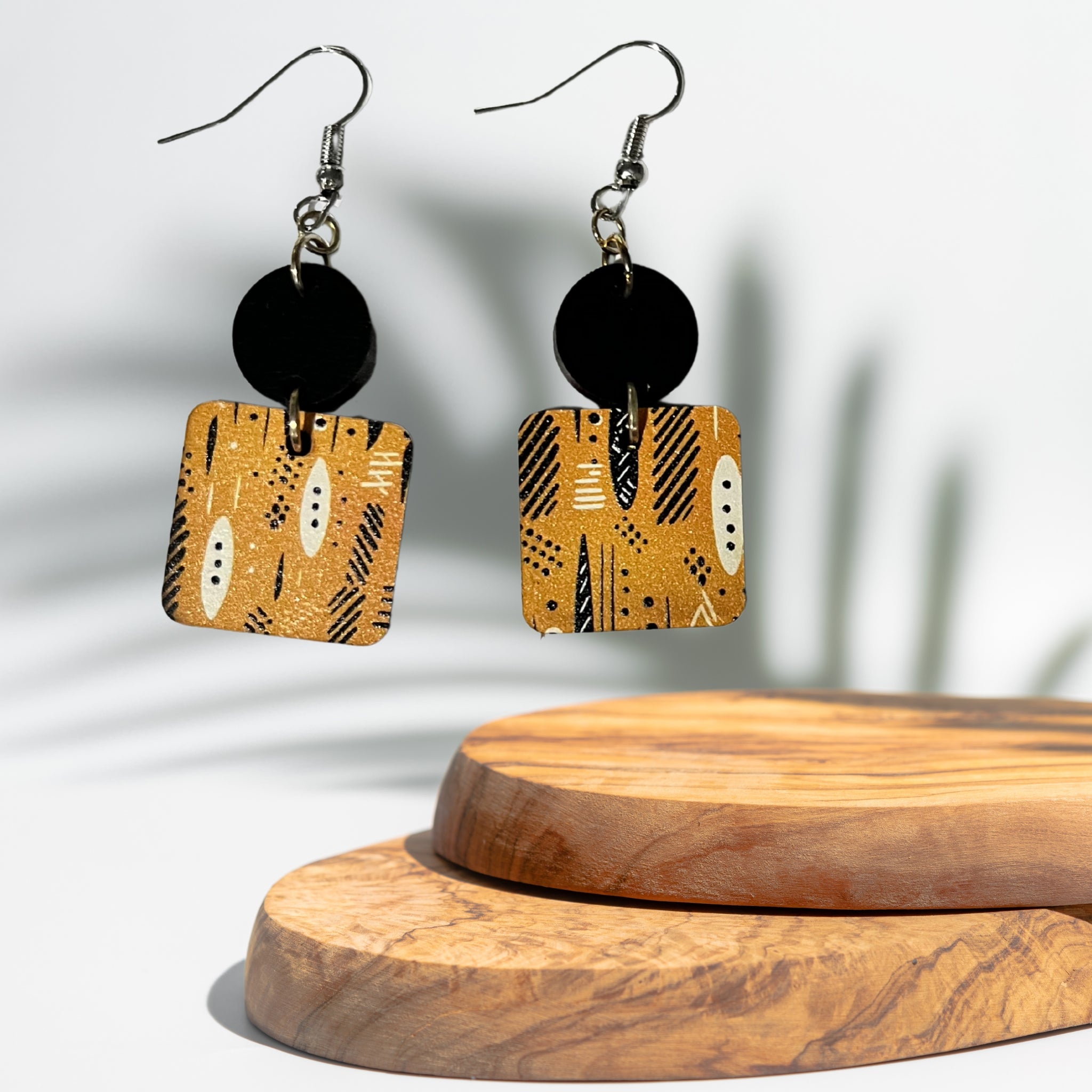 A pair of geometric drop earrings with black circular discs and square gold-toned charms featuring black and white tribal-style patterns, displayed on a wooden stand against a white background with soft leaf shadows.