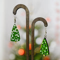 Two green tree-shaped dangle earrings with white and black geometric patterns, hanging from a dark wooden display stand against a softly blurred holiday background.