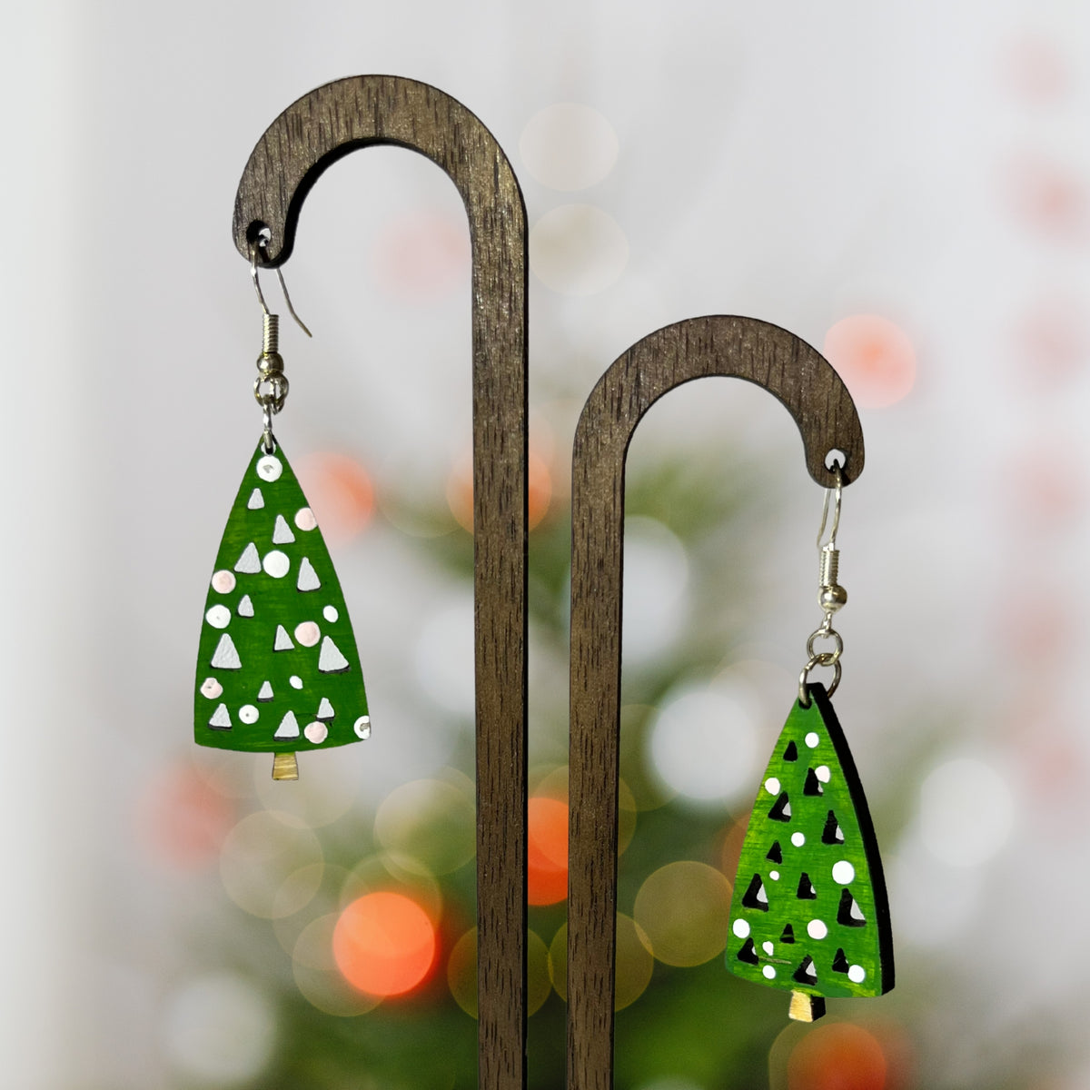 Two green tree-shaped dangle earrings with white and black geometric patterns, hanging from a dark wooden display stand against a softly blurred holiday background.