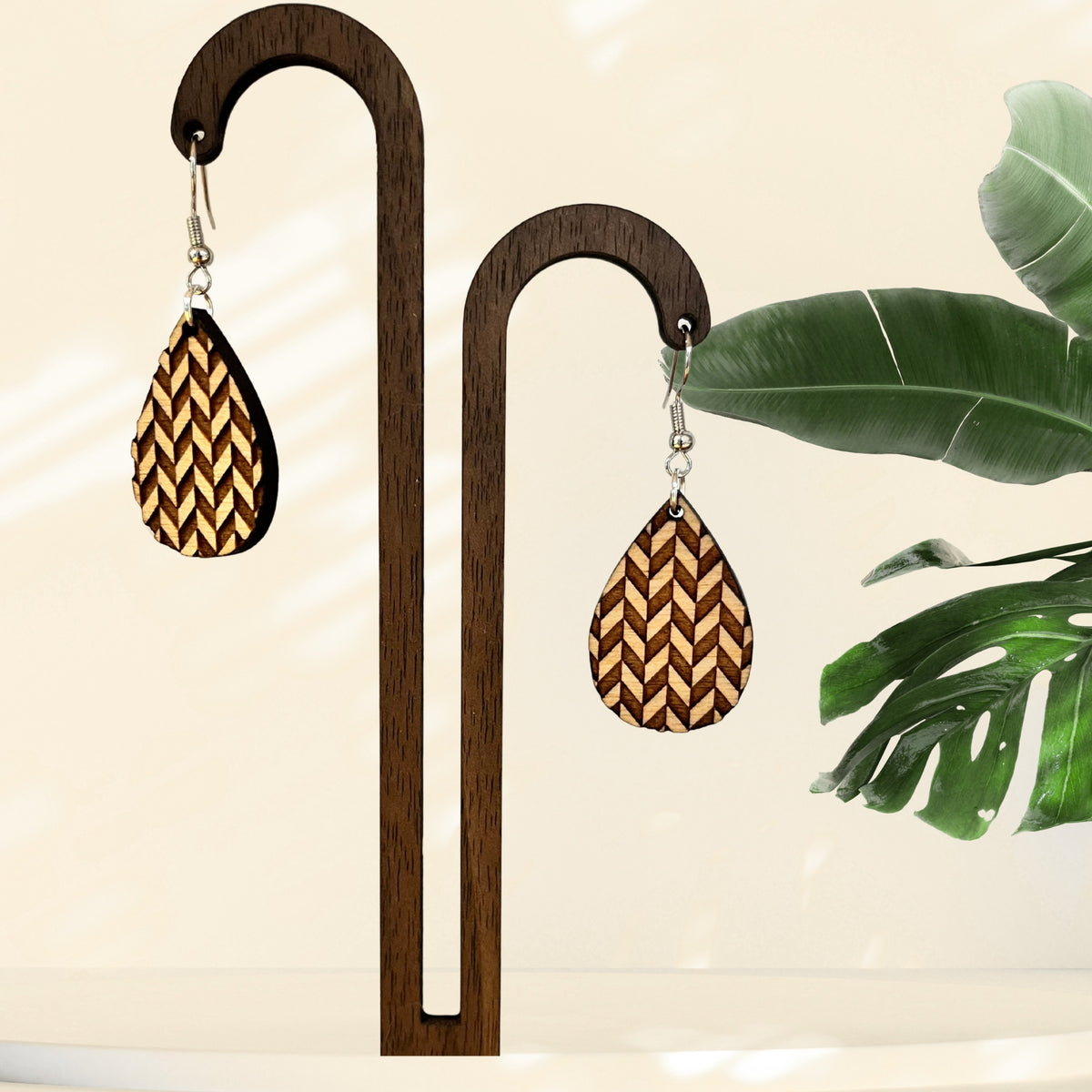 Teardrop-shaped wooden earrings with a laser-engraved chevron pattern in natural wood tones, displayed on a dark wood stand with tropical leaves in the background.