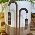 A pair of rectangular drop earrings with dark wood and black striped patterns, hanging from a wooden jewelry display stand in front of a white ceramic planter with green leaves.