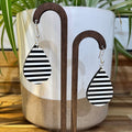 Black and white striped teardrop dangle earrings displayed on a wooden stand in front of a white ceramic planter with green leaves in the background.