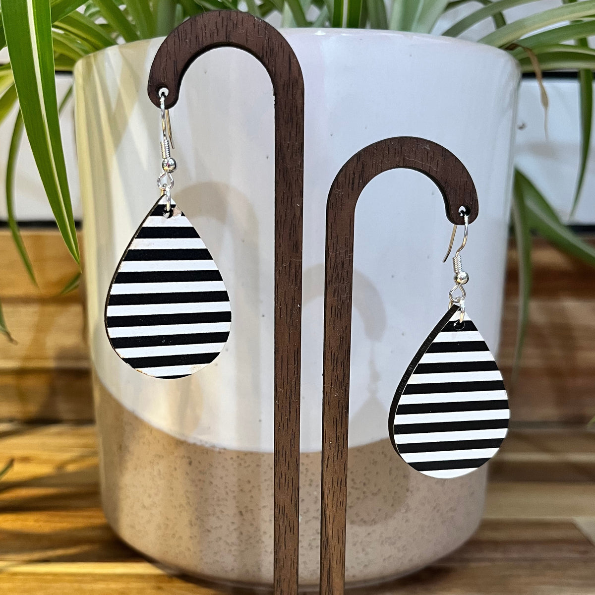 Black and white striped teardrop dangle earrings displayed on a wooden stand in front of a white ceramic planter with green leaves in the background.