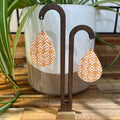 A pair of teardrop-shaped dangle earrings with a white chevron pattern on a tan background, hanging from a dark wood display stand in front of a white planter with green plant leaves.