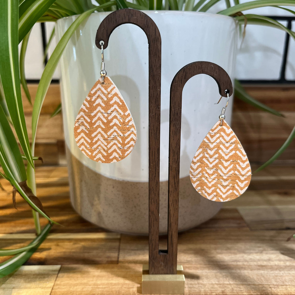 A pair of teardrop-shaped dangle earrings with a white chevron pattern on a tan background, hanging from a dark wood display stand in front of a white planter with green plant leaves.