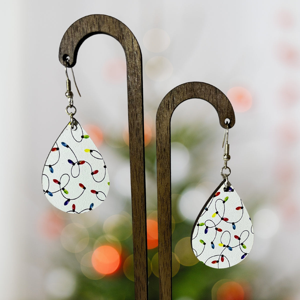 Teardrop-shaped white earrings printed with colorful Christmas string lights, hanging from a dark wooden display stand against a softly blurred holiday background.