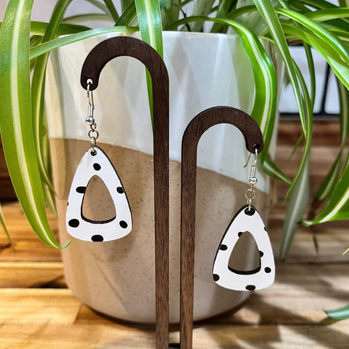 White triangle dangle earrings with black polka dots, displayed on a wooden stand in front of a potted spider plant.