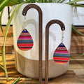 Teardrop-shaped dangle earrings with colorful horizontal striped fabric panels, displayed on a wooden stand in front of a white ceramic planter with green plant leaves.