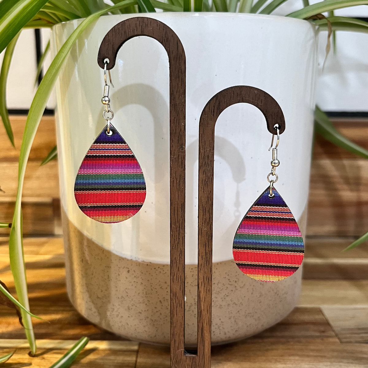 Teardrop-shaped dangle earrings with colorful horizontal striped fabric panels, displayed on a wooden stand in front of a white ceramic planter with green plant leaves.