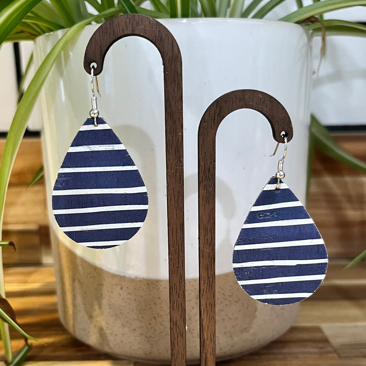 Teardrop-shaped wooden dangle earrings with navy blue and white horizontal stripes, displayed on a white ceramic planter with green plant leaves in the background.