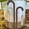 A pair of rectangular drop earrings with a light wood grain finish, displayed on a wooden stand in front of a white ceramic pot with green plant leaves.