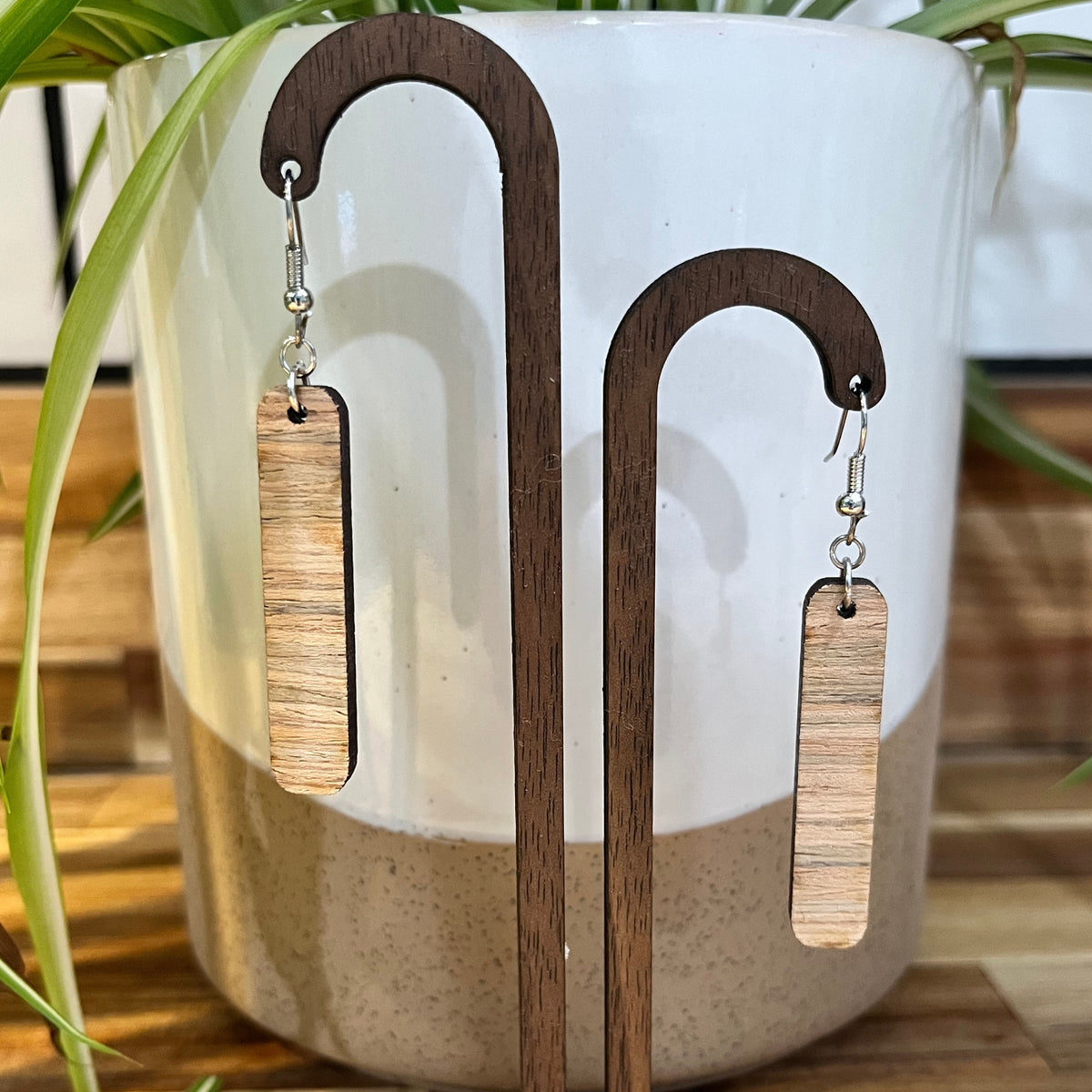 A pair of rectangular drop earrings with a light wood grain finish, displayed on a wooden stand in front of a white ceramic pot with green plant leaves.