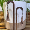 A pair of wooden dangle earrings with triangular pendant engraved with a pine tree, displayed on a ceramic plant pot on a wooden surface.