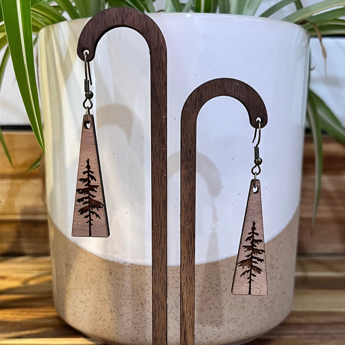 A pair of wooden dangle earrings with triangular pendant engraved with a pine tree, displayed on a ceramic plant pot on a wooden surface.