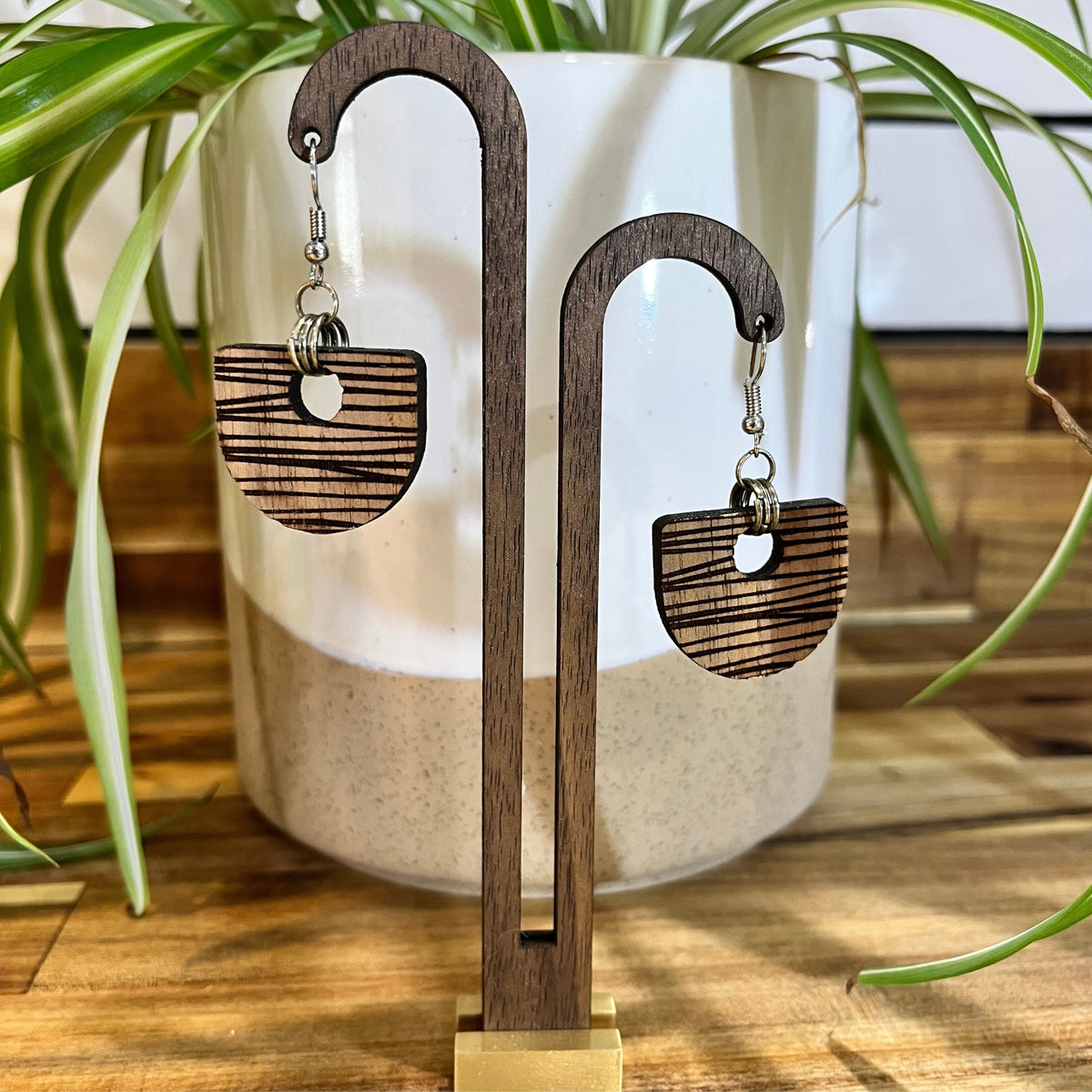 Wooden dangle earrings with a striped laser-burned finish, displayed on a wooden stand in front of a white ceramic planter with green plant leaves.