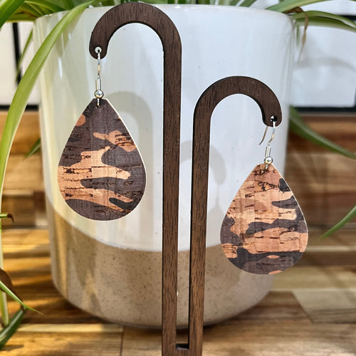 Teardrop-shaped wooden earrings with a brown and tan camouflage pattern, displayed on a dark wood stand in front of a white ceramic planter with green leaves.