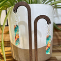 A pair of rectangular drop earrings with a colorful marbled pattern in teal, orange, and cream, hanging from gold hooks on a wooden display stand in front of a white ceramic planter.