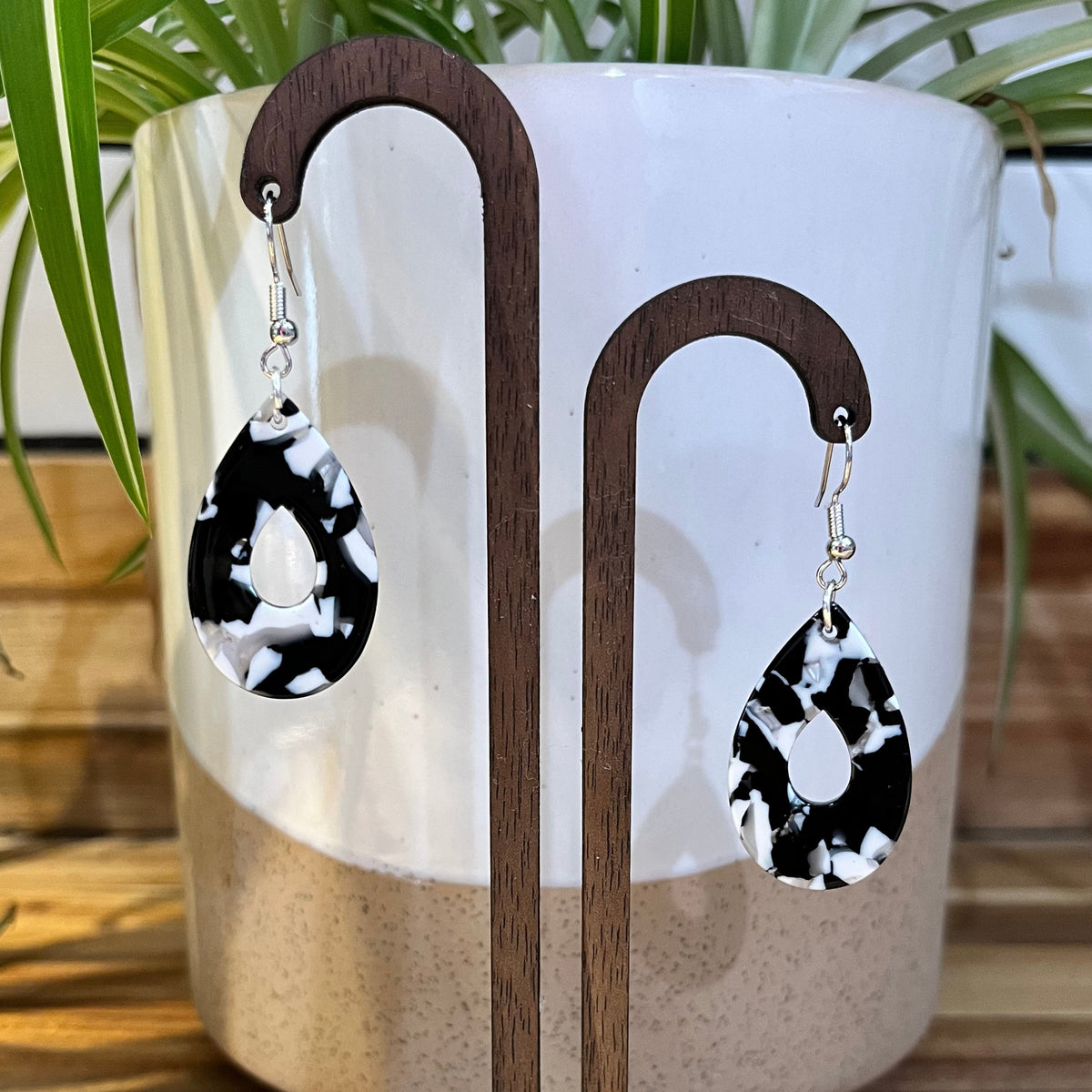 Teardrop-shaped dangle earrings with a black and white marbled pattern, hanging from silver fishhook wires on a wooden display stand in front of a white ceramic planter with green leaves.