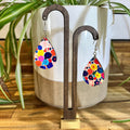 Teardrop-shaped dangle earrings with a colorful floral pattern, displayed on a dark wood stand in front of a white ceramic planter with green plant leaves.